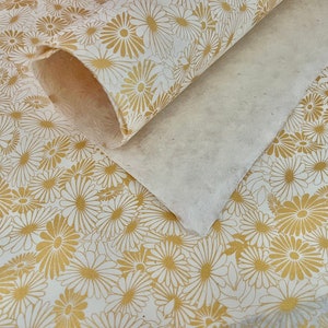 K&ouml;nnte beinhalten: Wei&szlig;es Geschenkpapier mit goldenem Blumenmuster. Das Papier ist aufgerollt und die Kanten sind sichtbar.