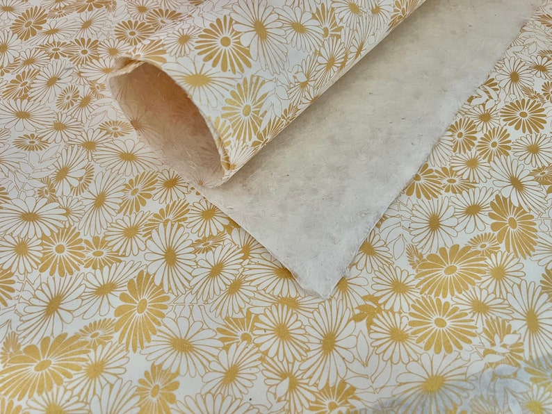 K&ouml;nnte beinhalten: Wei&szlig;es Geschenkpapier mit goldenem Blumenmuster. Das Papier ist aufgerollt und die Kanten sind sichtbar.