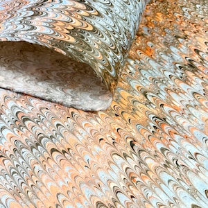 May include: A close-up of a rolled sheet of marbled paper. The paper features a repeating pattern of curved lines in shades of orange, black, brown, and light blue. The paper is likely used for decorative purposes or crafts.