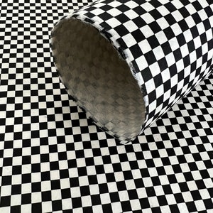 May include: A cylindrical tube covered in a black and white checkered pattern, resting on a flat surface featuring the same repeating black and white checkerboard design. The interior of the tube shows a textured, light-colored fibrous material.
