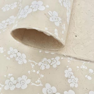 Puede incluir: Un rollo de papel beige con un patr&oacute;n floral blanco. El papel tiene una superficie texturizada y es perfecto para manualidades o envolver regalos.