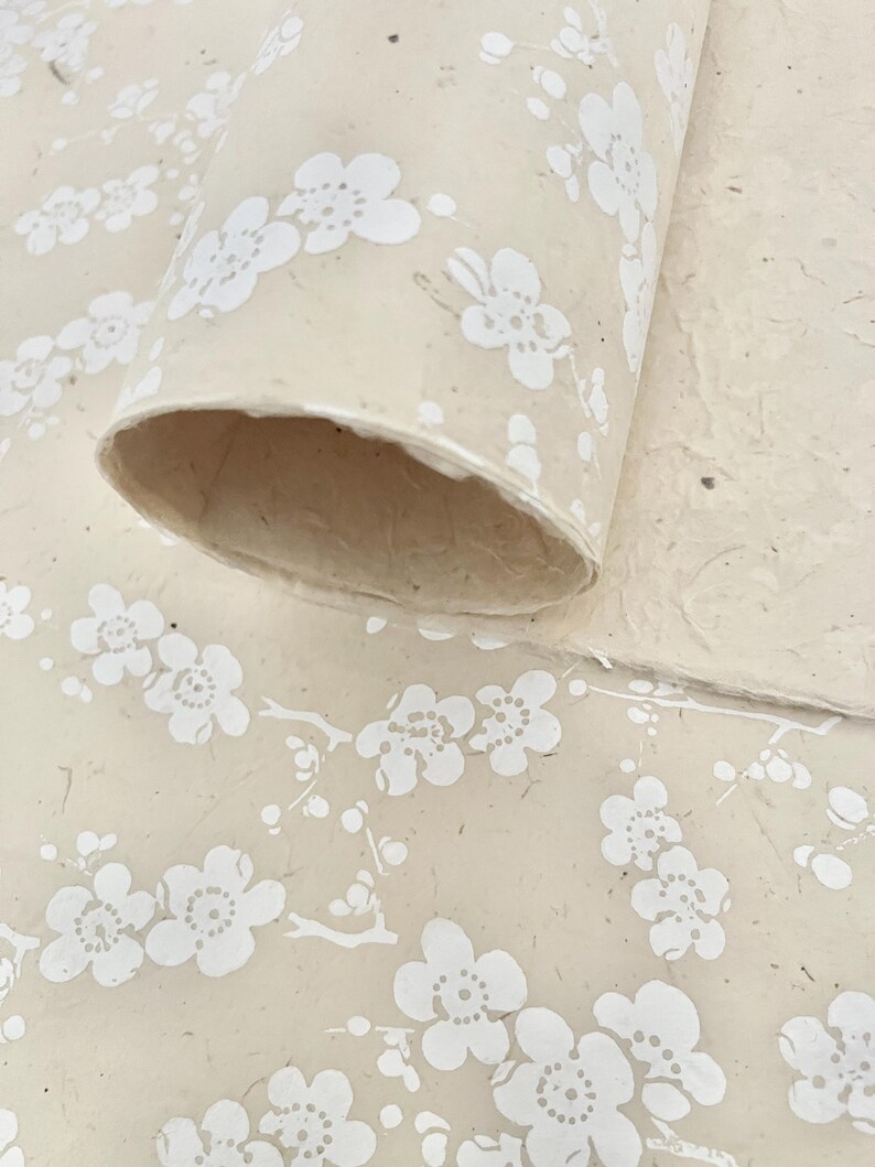 Puede incluir: Un rollo de papel beige con un patr&oacute;n floral blanco. El papel tiene una superficie texturizada y es perfecto para manualidades o envolver regalos.