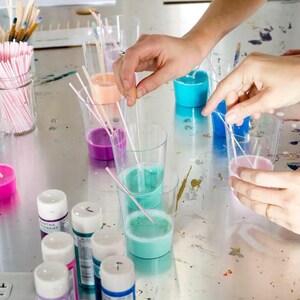 May include: Clear plastic cups filled with various shades of liquid paint including blue, pink, purple, and orange, each containing a thin white stirring straw. Several small paint bottles with white caps are arranged in the foreground.