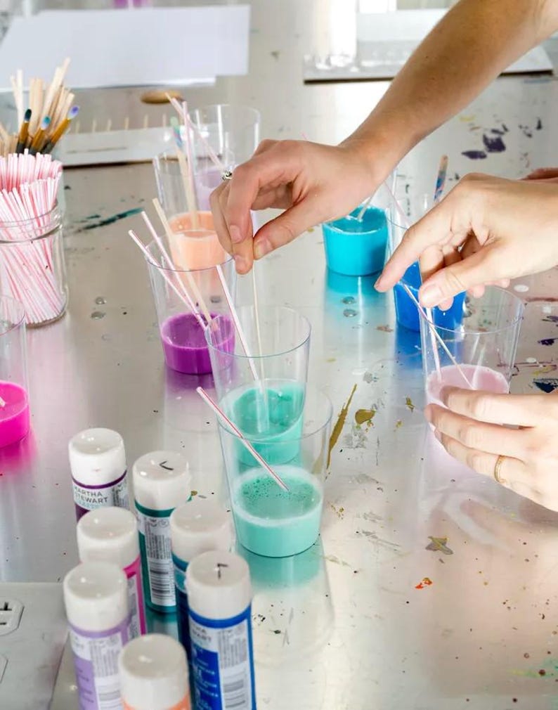 May include: Clear plastic cups filled with various shades of liquid paint including blue, pink, purple, and orange, each containing a thin white stirring straw. Several small paint bottles with white caps are arranged in the foreground.