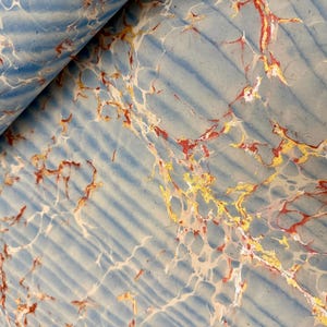 May include: Close-up of a sheet of paper with a marbled design. The paper has a light blue background with darker blue diagonal stripes. Veins of red, gold, and white create a marbled effect. The paper is partially rolled.
