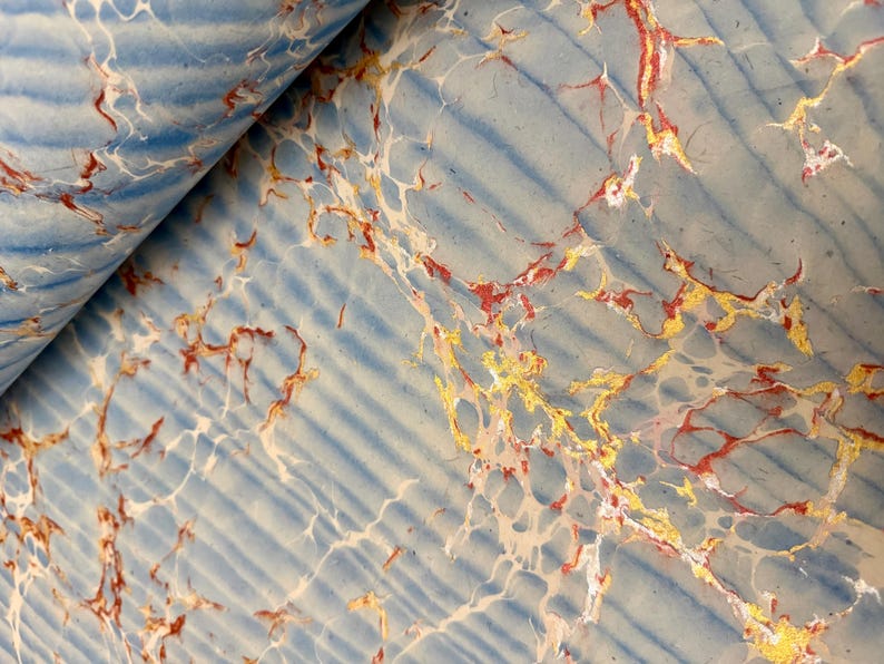 May include: Close-up of a sheet of paper with a marbled design. The paper has a light blue background with darker blue diagonal stripes. Veins of red, gold, and white create a marbled effect. The paper is partially rolled.