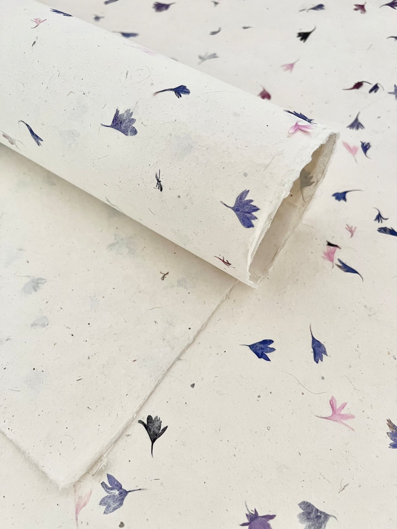 May include: A roll of white paper with a scattered pattern of small, dried blue and pink flowers. The paper has a textured, handmade look.