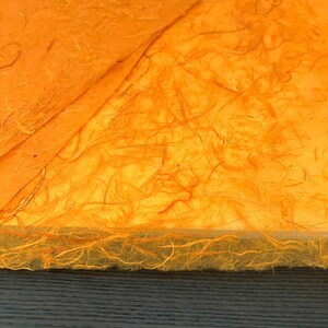 May include: Sheets of orange handmade paper with a fibrous, textured surface. The paper features visible organic plant fibers embedded throughout the material, creating a translucent and uneven appearance with a rough, deckled edge along the bottom.