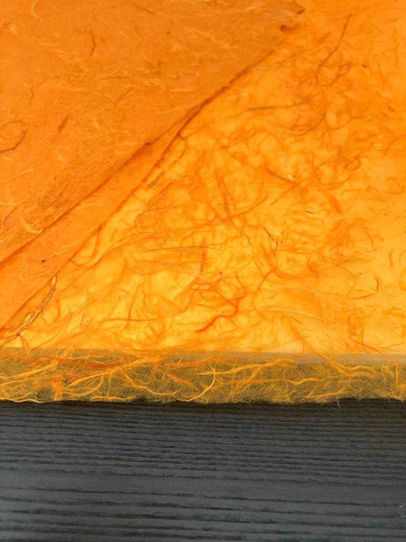 May include: Sheets of orange handmade paper with a fibrous, textured surface. The paper features visible organic plant fibers embedded throughout the material, creating a translucent and uneven appearance with a rough, deckled edge along the bottom.