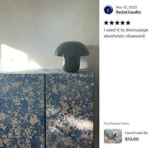 May include: A rectangular credenza covered in blue and white marbled paper with a swirling pattern, featuring a dark green mushroom-shaped lamp placed on top. The surface displays a complex mix of blue tones and white circular shapes.