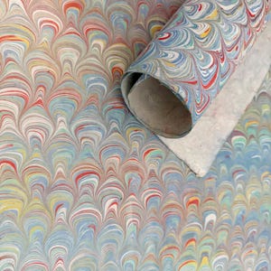 May include: A roll of paper featuring a marbled pattern with repeating wave-like shapes in shades of light blue, red, yellow, and white. The paper has a smooth surface texture and is coiled into a cylindrical shape, revealing a plain white underside.