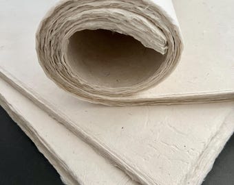 May include: A roll of off-white, textured handmade paper resting on top of several flat, stacked sheets of the same material. The paper features a visible fibrous composition with uneven, deckled edges and a thick, organic, and porous surface texture.