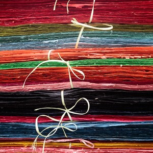 May include: A stack of colorful handmade paper sheets, bound with natural twine. The paper is in various shades of red, orange, green, blue, and black. The texture of the paper is visible, suggesting a craft or art supply.