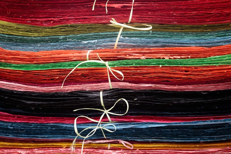 May include: A stack of colorful handmade paper sheets, bound with natural twine. The paper is in various shades of red, orange, green, blue, and black. The texture of the paper is visible, suggesting a craft or art supply.