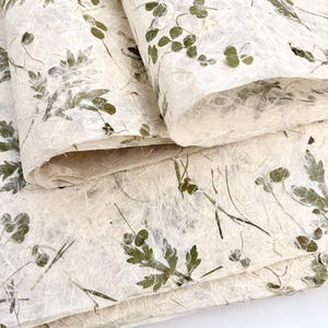 May include: Close-up of handmade paper with embedded green leaves and stems. The paper has a textured, fibrous appearance with a natural, off-white color. The leaves and stems are arranged throughout the paper, creating a botanical design.