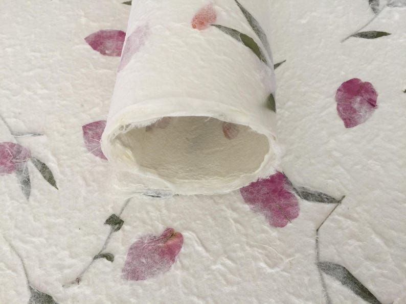 May include: A rolled sheet of handmade paper with a textured surface. The paper is white with pink floral accents and green leaves. The paper is likely used for crafts or decorative purposes.