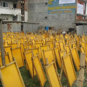 May include: A field of yellow paper drying on wooden frames. The frames are propped up on wooden supports, arranged in rows on the grass. Buildings and a sign are visible in the background.