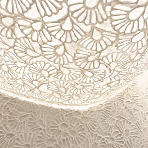 May include: A white bowl featuring a repeating cutout pattern of stylized flower petals. The bowl has a translucent appearance, and the same floral motif is visible on the surface beneath it, creating a layered effect of white shapes.