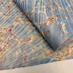 May include: A sheet of marbled paper with a light blue background and a ribbed texture. The paper features intricate patterns of white, gold, and red veins, creating a unique and artistic design. The paper is folded over, revealing its layered construction.