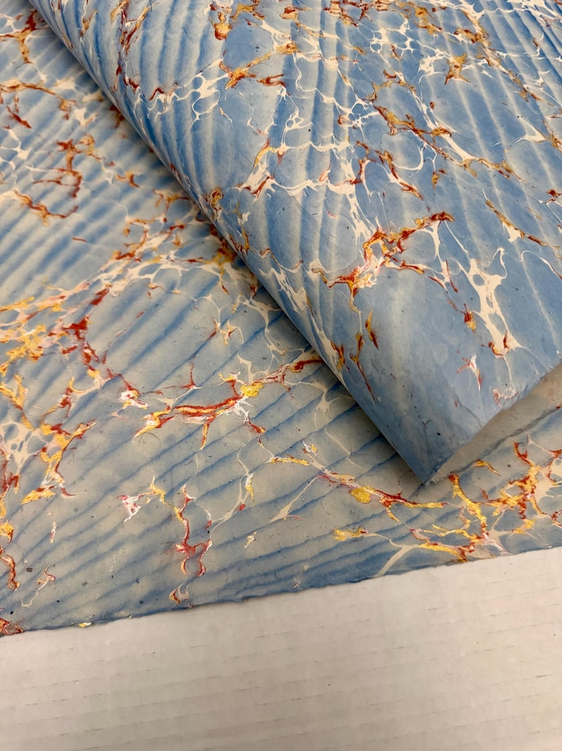 May include: A sheet of marbled paper with a light blue background and a ribbed texture. The paper features intricate patterns of white, gold, and red veins, creating a unique and artistic design. The paper is folded over, revealing its layered construction.