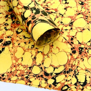 May include: A rolled sheet of marbled paper with a yellow base, featuring black and reddish-brown organic patterns. The paper is partially rolled, revealing the interior. The design is reminiscent of natural stone or abstract art.