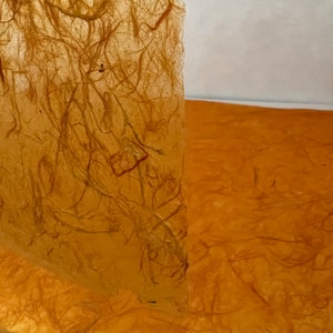May include: A sheet of translucent, amber-colored handmade paper featuring a visible network of embedded organic fibers. The texture is fibrous and uneven, with a warm, golden-brown hue that allows light to pass through the delicate, thin material.