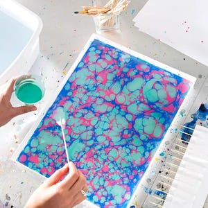 May include: A rectangular paper with a marbled design in shades of blue, pink, and teal is being created. The process involves applying paint to the surface with a stick. A cup of teal paint and a container of water are nearby.