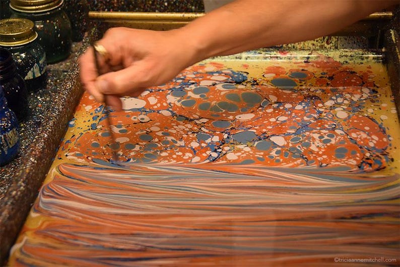 May include: A rectangular tray filled with liquid containing swirling patterns of orange, blue, and white paint. A thin tool is being used to manipulate the colors on the surface, creating marbled designs with circular and elongated streaks.