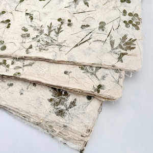 Puede incluir: Una pila de hojas de papel hechas a mano con elementos botánicos incrustados. El papel tiene un color blanquecino natural con inclusiones vegetales verdes y marrones, creando una apariencia texturizada y orgánica. Las hojas están superpuestas.