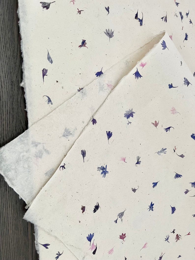May include: A sheet of handmade paper with a scattered pattern of pressed blue, pink, and purple flowers.