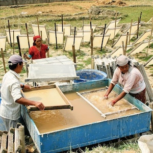 May include: Three people working in a paper making workshop. Two people are using screens to lift wet paper pulp from a large blue tub. The third person is standing next to a tub and holding a screen with a sheet of paper pulp. There are many wooden frames drying in the background.