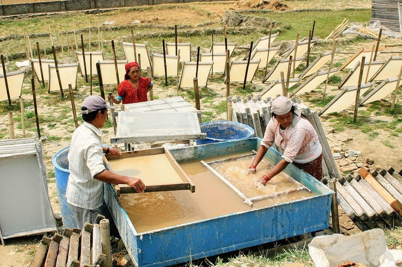 May include: Three people working in a paper making workshop. Two people are using screens to lift wet paper pulp from a large blue tub. The third person is standing next to a tub and holding a screen with a sheet of paper pulp. There are many wooden frames drying in the background.