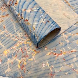 May include: A rolled sheet of marbled paper with a blue and white background. The paper has a pattern of blue lines and veins of red and gold. The paper is partially rolled, revealing the inside of the roll.