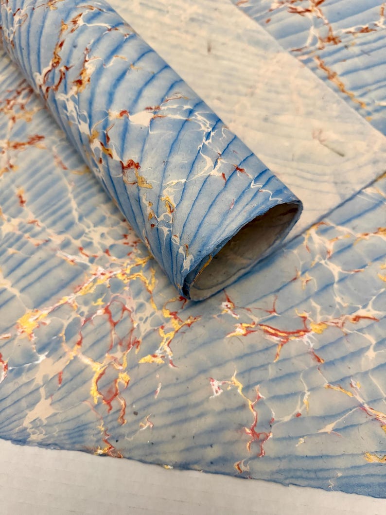 May include: A rolled sheet of marbled paper with a blue and white background. The paper has a pattern of blue lines and veins of red and gold. The paper is partially rolled, revealing the inside of the roll.
