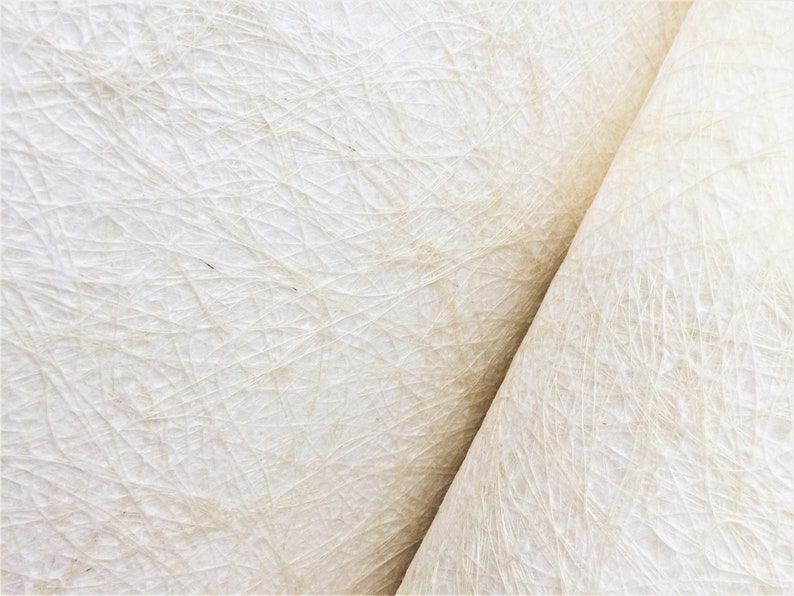 Op de afbeelding: Close-up van een wit, getextureerd papier met een subtiel geweven patroon. Het papier is doormidden gevouwen, waardoor een diagonale lijn over de afbeelding loopt.