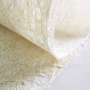 Op de afbeelding: Close-up van een vel wit handgeschept papier met een textuur, gemaakt van vezels. Het papier is dubbelgevouwen, waardoor de delicate vezels zichtbaar worden die de structuur vormen.