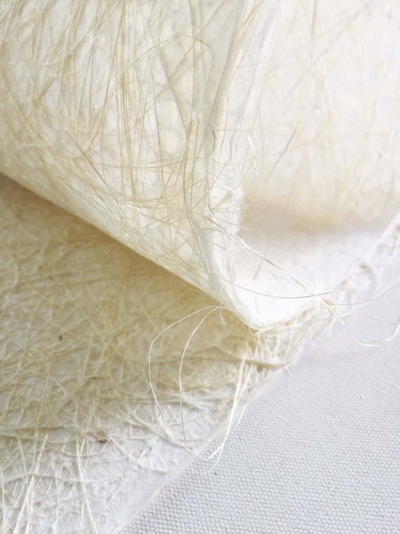 Op de afbeelding: Close-up van een vel wit handgeschept papier met een textuur, gemaakt van vezels. Het papier is dubbelgevouwen, waardoor de delicate vezels zichtbaar worden die de structuur vormen.