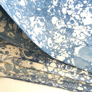 May include: A stack of sheets of paper featuring a marbled pattern in shades of blue and white. The design consists of various sized circular and organic shapes created with blue ink on a white background, layered to show the texture of the paper.