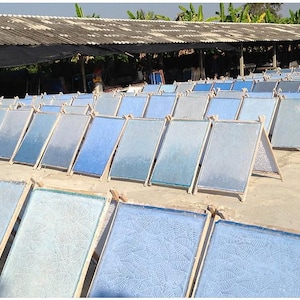May include: A collection of rectangular, light blue paper sheets drying outdoors on wooden frames. The sheets vary in shade, with some displaying a subtle leaf pattern. A covered structure and greenery are visible in the background.
