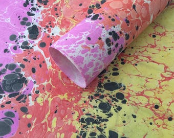 May include: A roll of marbled paper with a vibrant pattern. The design features pink, red, yellow, and black swirls and spots. The paper is rolled up, revealing the inside edge. This decorative paper is suitable for crafts and art projects.