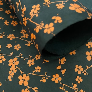 May include: A roll of black paper featuring a repeating pattern of orange cherry blossom branches. The paper has a textured surface and is partially unrolled, displaying the floral design across the dark background in a consistent, repeating layout.