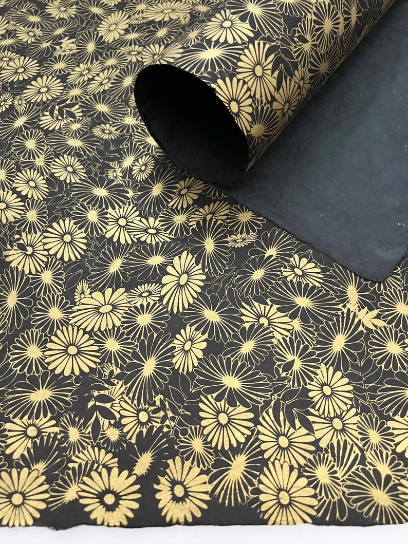 K&ouml;nnte beinhalten: Schwarzes Geschenkpapier mit goldenem Blumenmuster. Das Papier ist aufgerollt und das Muster ist auf der Au&szlig;enseite der Rolle sichtbar.