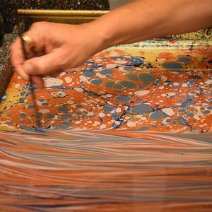 May include: A hand uses a tool to manipulate colorful inks on a liquid surface, creating a marbled effect. The colors include orange, blue, white, and yellow, with a striped pattern at the bottom. Jars of paint are visible in the background, suggesting a craft project.