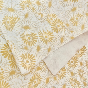 K&ouml;nnte beinhalten: Wei&szlig;es Geschenkpapier mit einem sich wiederholenden Muster aus goldenen G&auml;nsebl&uuml;mchen.