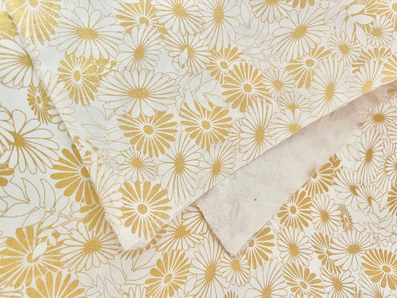K&ouml;nnte beinhalten: Wei&szlig;es Geschenkpapier mit einem sich wiederholenden Muster aus goldenen G&auml;nsebl&uuml;mchen.