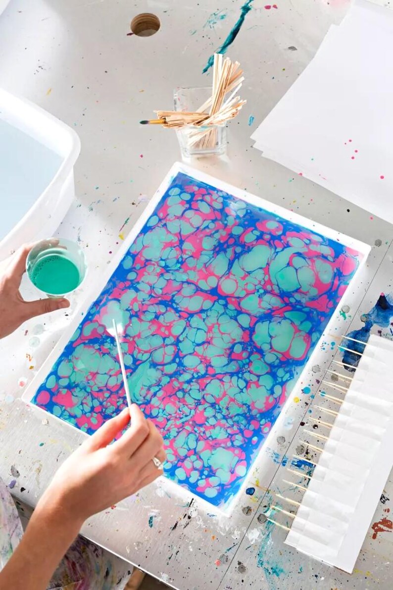 May include: A rectangular white tray filled with water and a swirling marbled pattern of bright pink, turquoise, and deep blue paint. A thin white stylus is held over the surface, while a small cup of turquoise paint sits nearby.