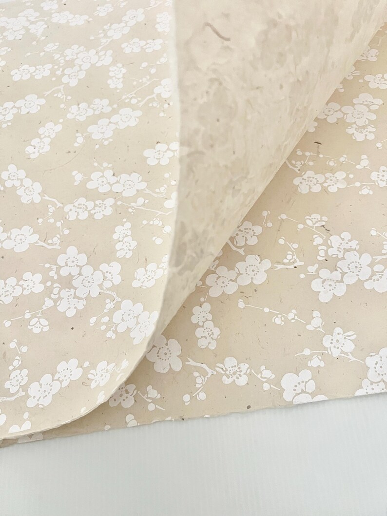 Puede incluir: Una hoja de papel beige con un patr&oacute;n floral blanco. El papel tiene una superficie texturizada y est&aacute; doblado por la mitad.