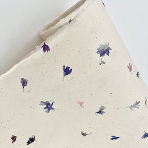 May include: A sheet of handmade paper with a delicate pattern of pressed purple and pink flowers. The paper has a natural, off-white color and a slightly textured surface.
