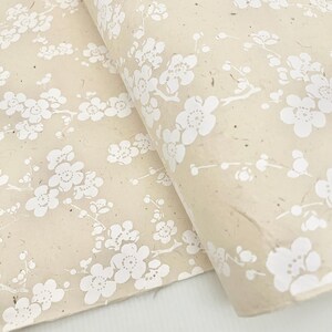 Puede incluir: Una hoja de papel de regalo beige con un patr&oacute;n floral blanco. El papel tiene un aspecto texturizado y hecho a mano.