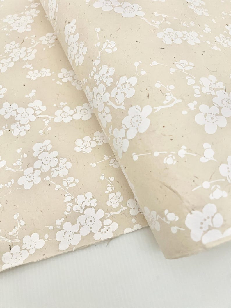 Puede incluir: Una hoja de papel de regalo beige con un patr&oacute;n floral blanco. El papel tiene un aspecto texturizado y hecho a mano.